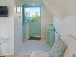 Un couloir étroit avec une porte donnant sur un balcon, une petite kitchenette équipée d’un évier et d’un plan de travail sur lequel se trouve un saladier, et un canapé agrémenté de coussins à motifs.