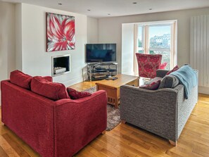 A living room with a red sofa, grey sofa, wooden coffee table, flat-screen TV, fireplace, and large windows. - 1 Richmond Place - UK51155 (St Ives)