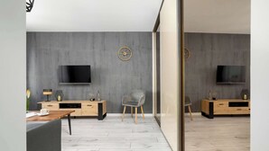Een moderne woonkamer met een grote flatscreen televisie aan de muur, een houten salontafel, een grijze bank en een grijze fauteuil.