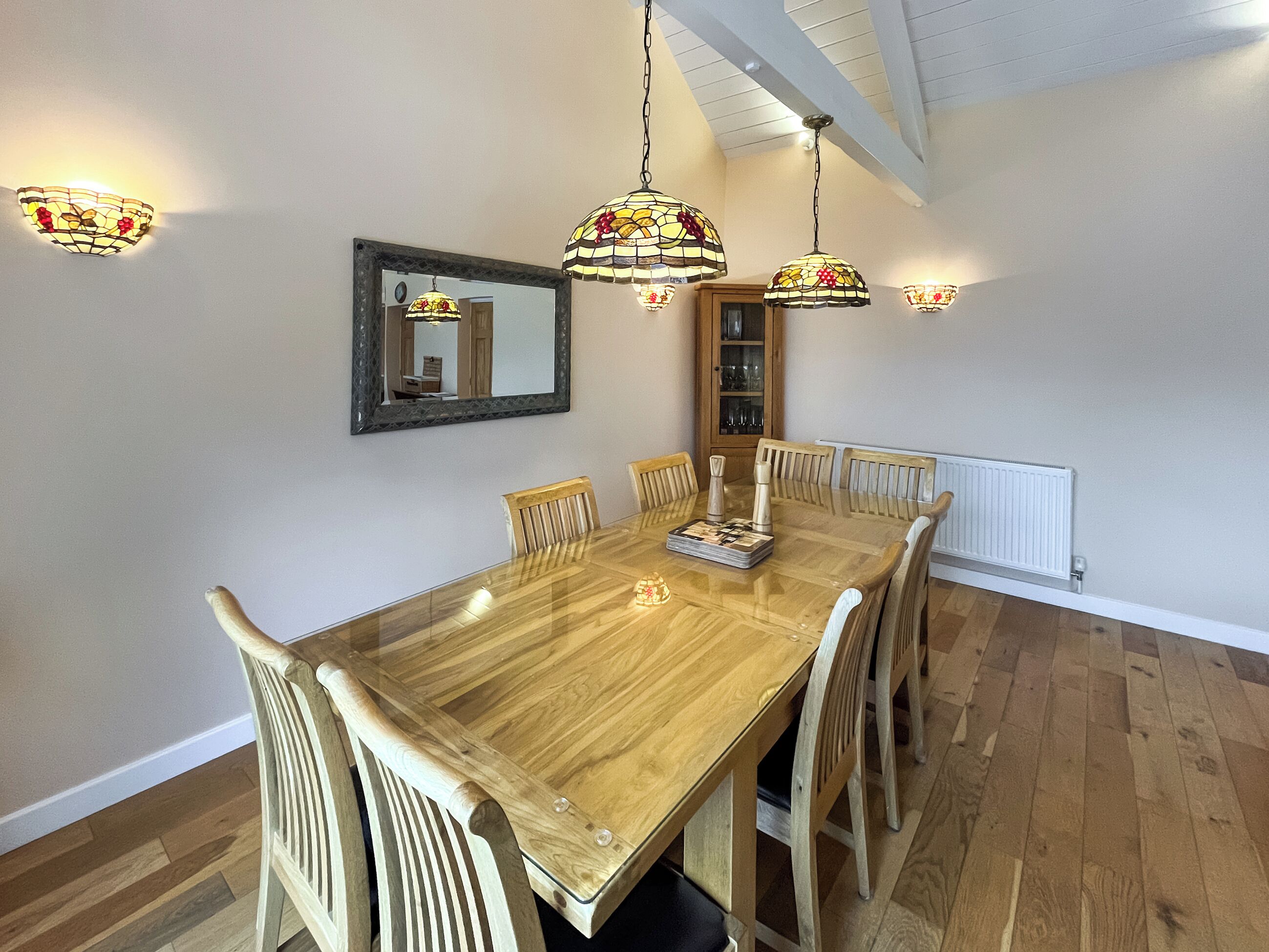 A dining area with a wooden table, six chairs, hanging stained glass lamps, a mirror, and a cabinet with glass doors.