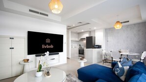 Un salon moderne doté d’un téléviseur à écran plat, d’une table basse en verre, de canapés bleus et d’un coin repas avec une table et des chaises blanches.