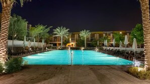 Un espace piscine nocturne avec des chaises longues et des parasols, entouré de palmiers et d’un bâtiment doté de nombreux balcons.