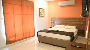 A hotel room with a bed, a nightstand, a remote control, a picture on the wall, and an air conditioning unit.