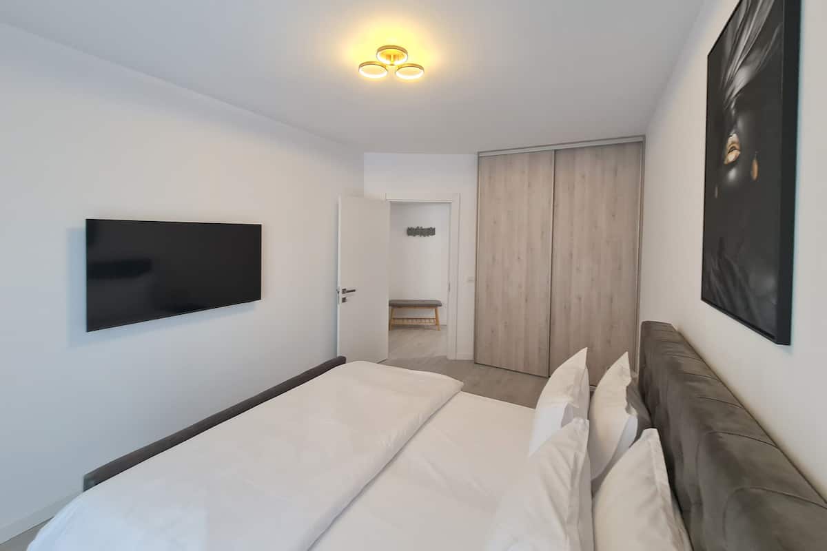 A modern bedroom with a bed, a flat-screen TV, a wardrobe, and a bench in the corner.