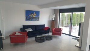 Una sala de estar moderna con un sofá azul oscuro, sillones rojos y dos otomanas. Un cuadro de un edificio azul adorna la pared, y hay un ventanal con vista a árboles.