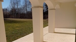 En åben patio med hvide søjler og udsigt over en græsplæne og træer.