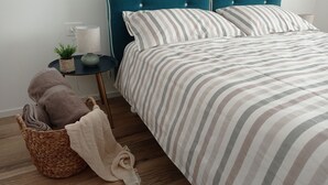 En pænt redt seng med stribet sengetøj, et teal-farvet polstret sengegavl og et natbord med en lampe og en lille plante.