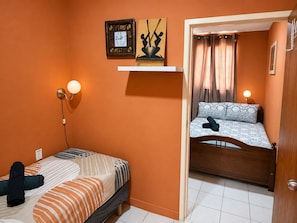 A compact room with a bed, a wall-mounted clock, a shelf with decorative items, and a door leading to another room. - Casa Andries – Centrally Located and Comfortable in Curaçao   (Willemstad)