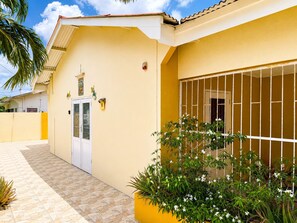 A yellow building with a gated entrance, a tiled patio, and a small garden with white flowers. - Casa Andries – Centrally Located and Comfortable in Curaçao   (Willemstad)