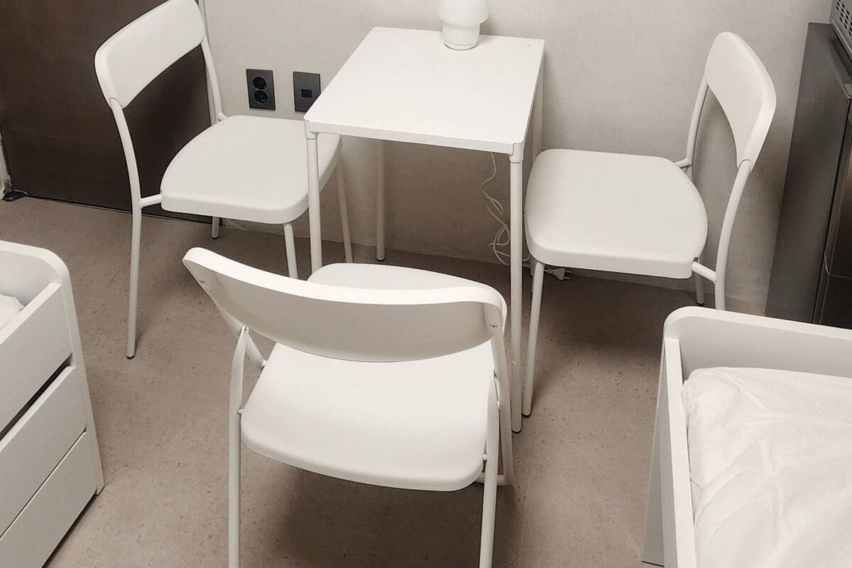 A small, clean room with a white desk, two chairs, a small table, and a bed.