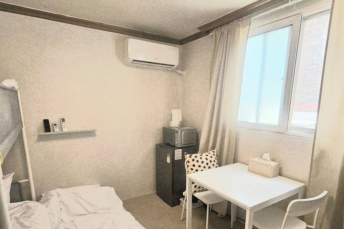 A hotel room with a bed, a desk, a chair, a small refrigerator, an air conditioning unit, and a window with curtains.
