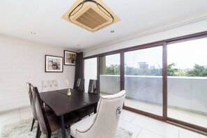 A dining area with a dark wooden table and chairs, a ceiling fan, and a large window with a city view. - Modern 3bdr Apt at Villaggio - Rooftop Pool & Skybar (Accra)