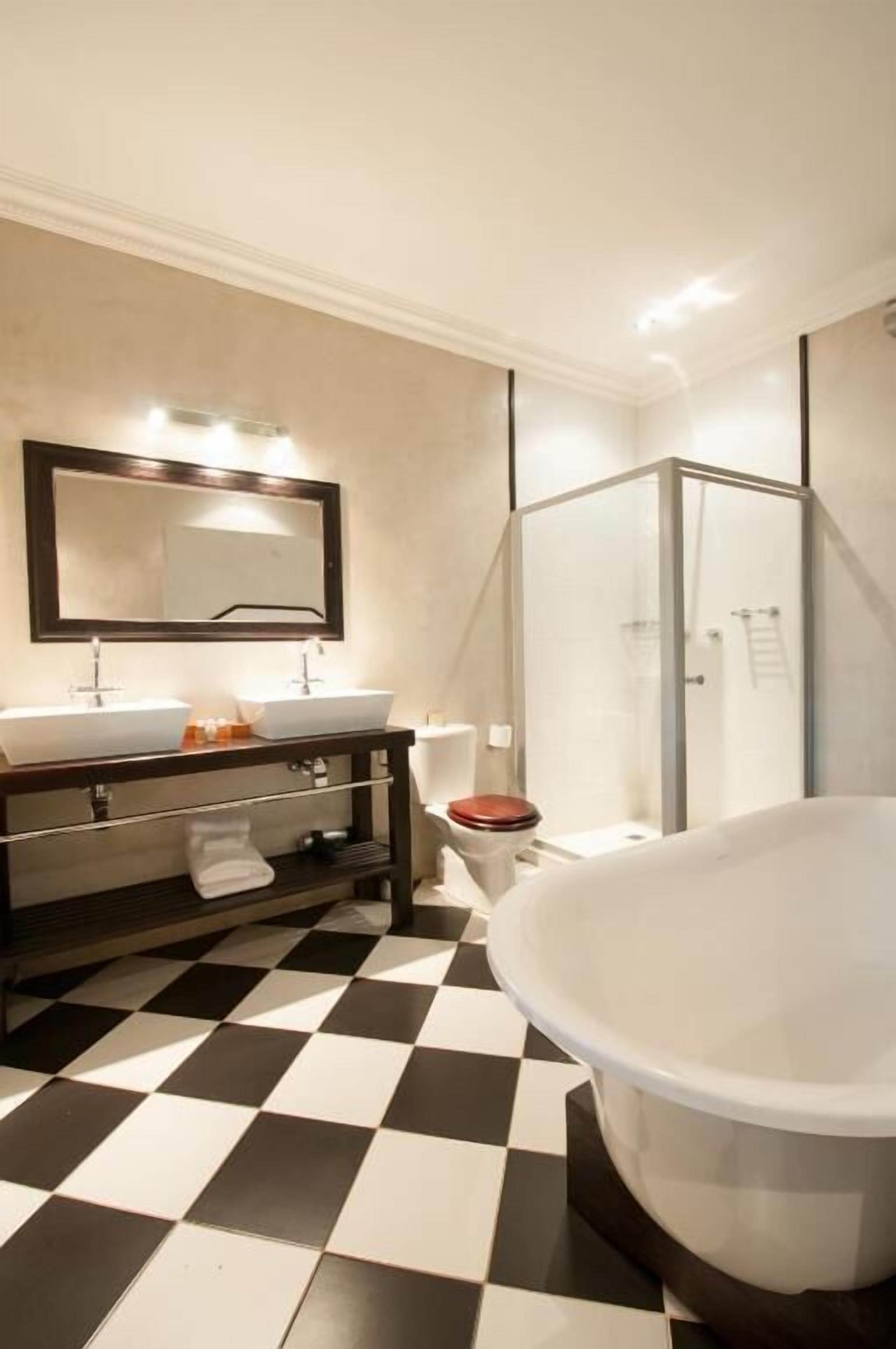 Un baño con piso a cuadros, una bañera grande, cabina de ducha de vidrio y tocador con espejo y dos lavamanos.