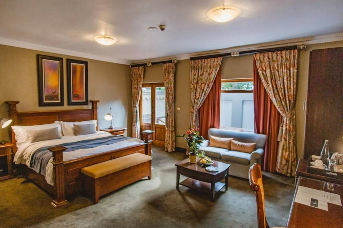 A hotel room with a large bed, a desk, a chair, and a sofa. There are curtains on the windows and a plant on the desk.