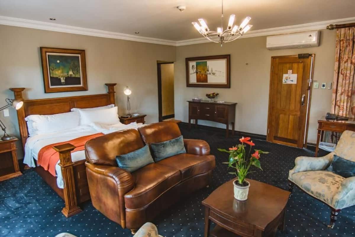 A hotel room with a bed, a leather sofa, a coffee table, a side table, and a chair. There are two paintings on the walls and a door with a sign.