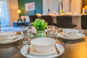 Une table à manger dressée avec des assiettes, des bols, des verres et des couverts, et un vase rempli de feuillage vert placé au centre.