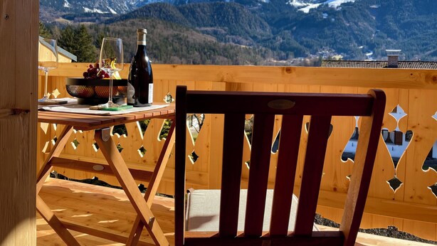 Una terraza de madera con una mesa dispuesta para dos personas, que incluye una botella de vino y un bol de uvas. Una silla se encuentra frente a la mesa. La terraza ofrece vistas a montañas nevadas bajo un cielo azul despejado.