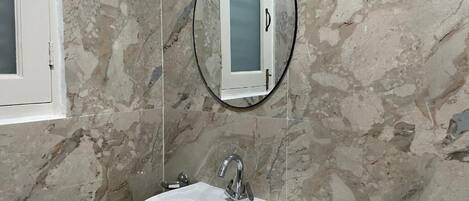 Un baño con pared de mármol, un lavamanos blanco, un espejo y una ventana.