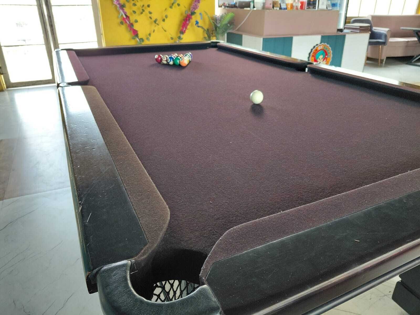 Una mesa de billar con una bola y taco, una pared amarilla con estampado floral, y un área de descanso con sofá y sillas.