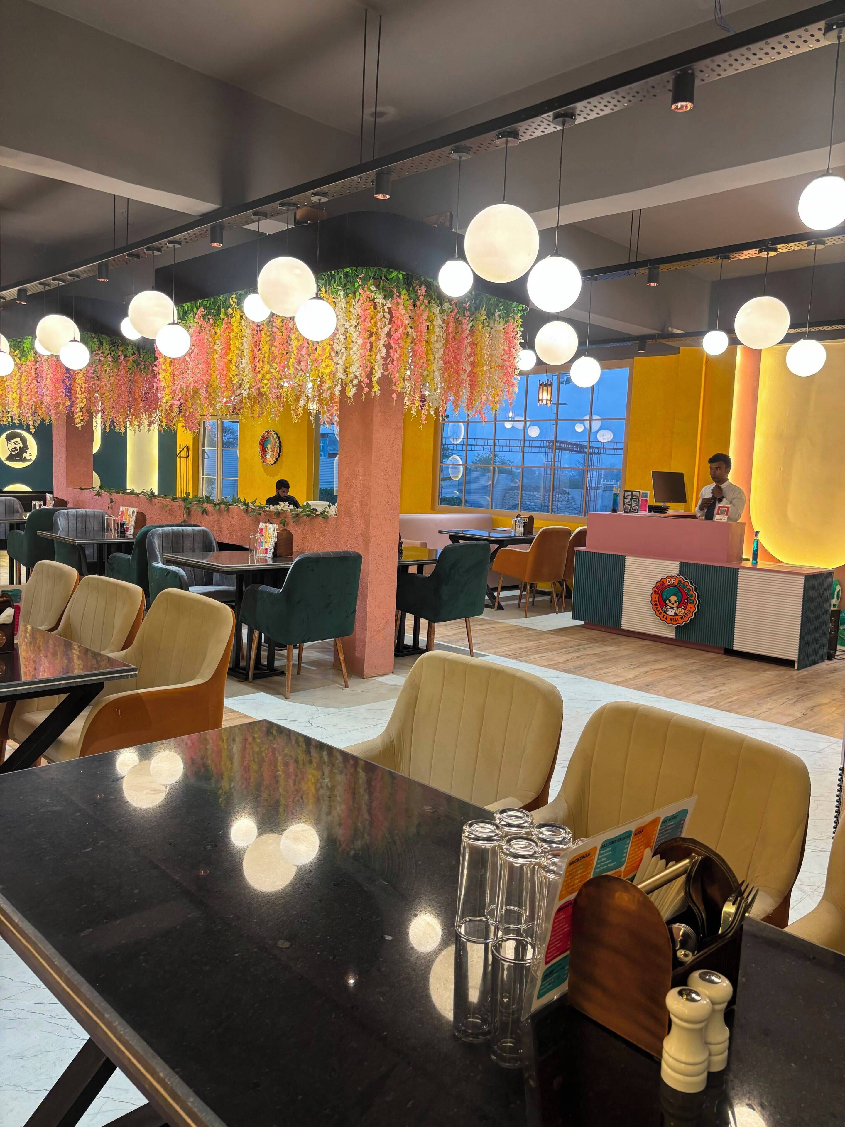 A modern restaurant interior with hanging floral decorations, a counter with a staff member, and multiple seating areas.