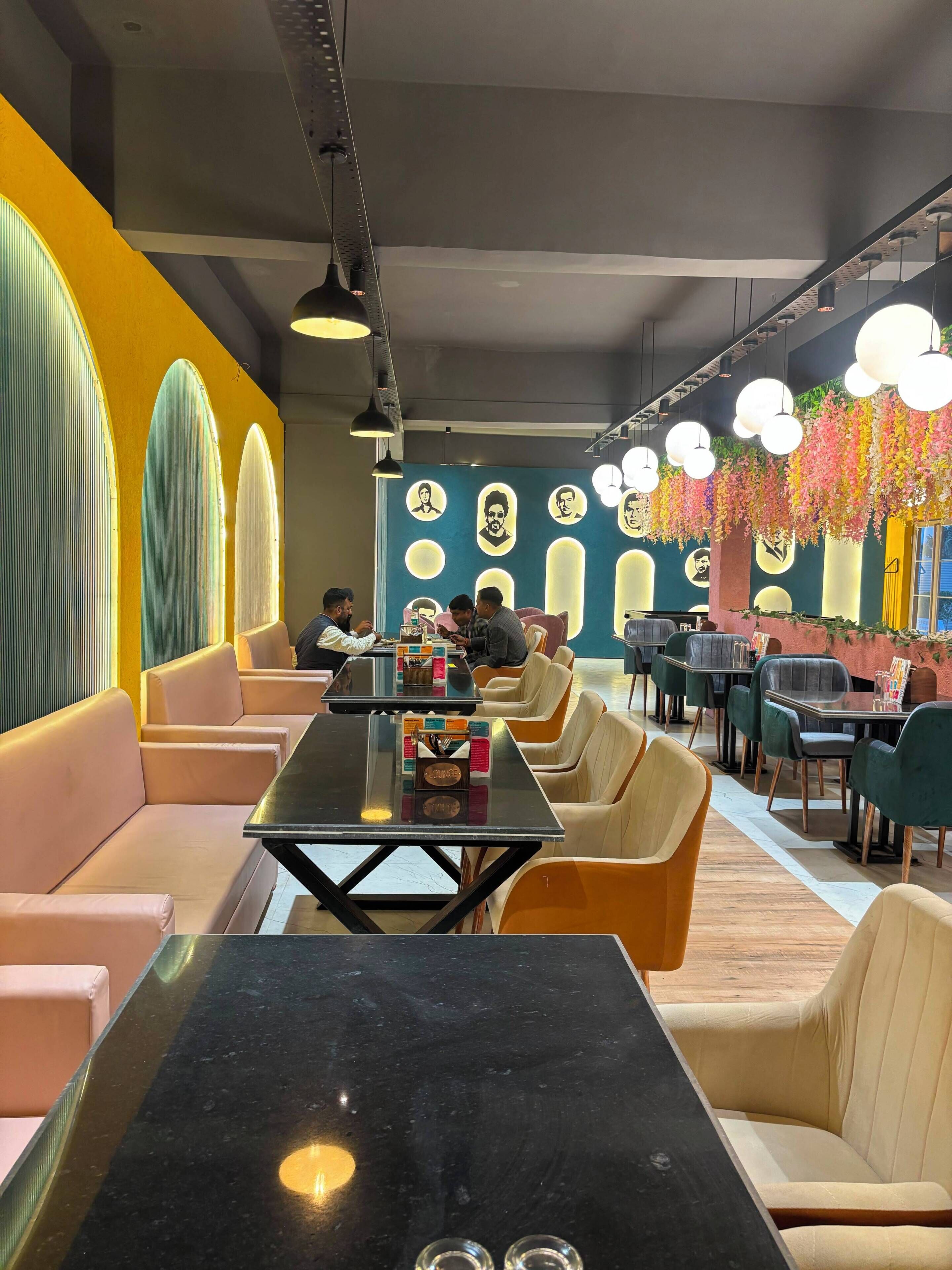 Et moderne restaurantinteriør med høje borde og polstrede siddepladser, en levende væg med cirkulære motiver, og hængende blomsterdekorationer.