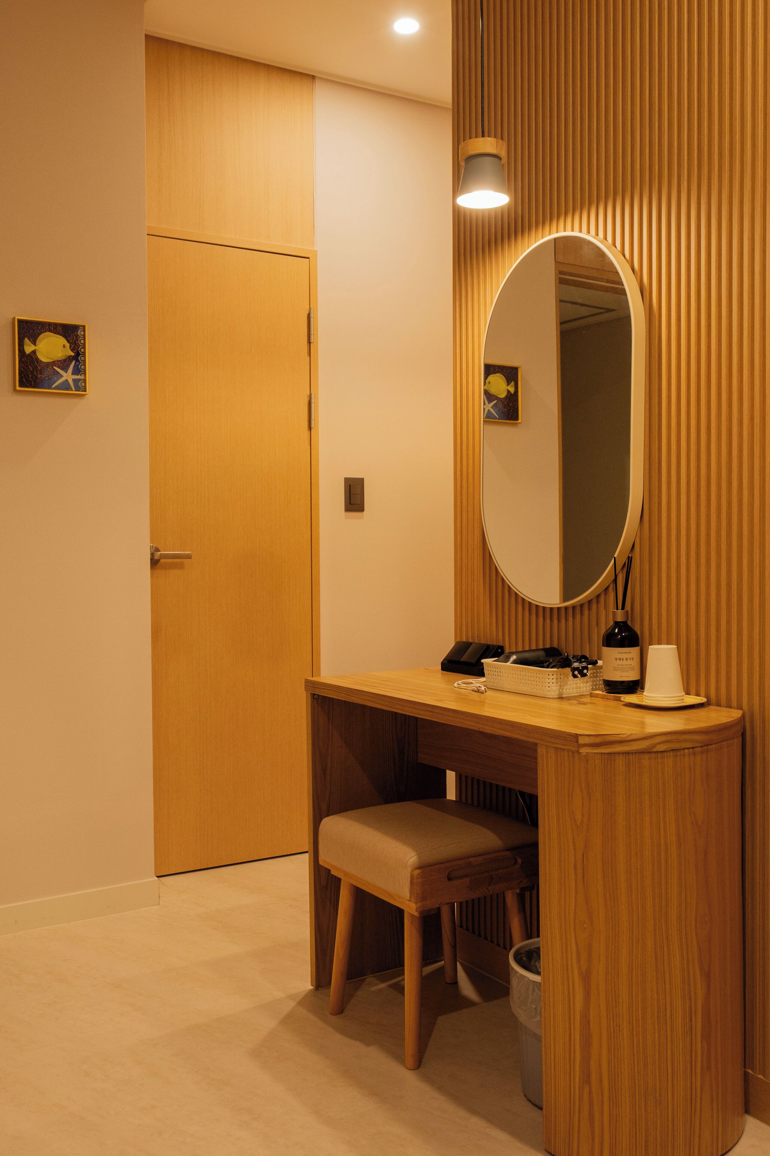 A hotel room with a wooden vanity, a mirror, a desk, a chair, a waste bin, and a door.