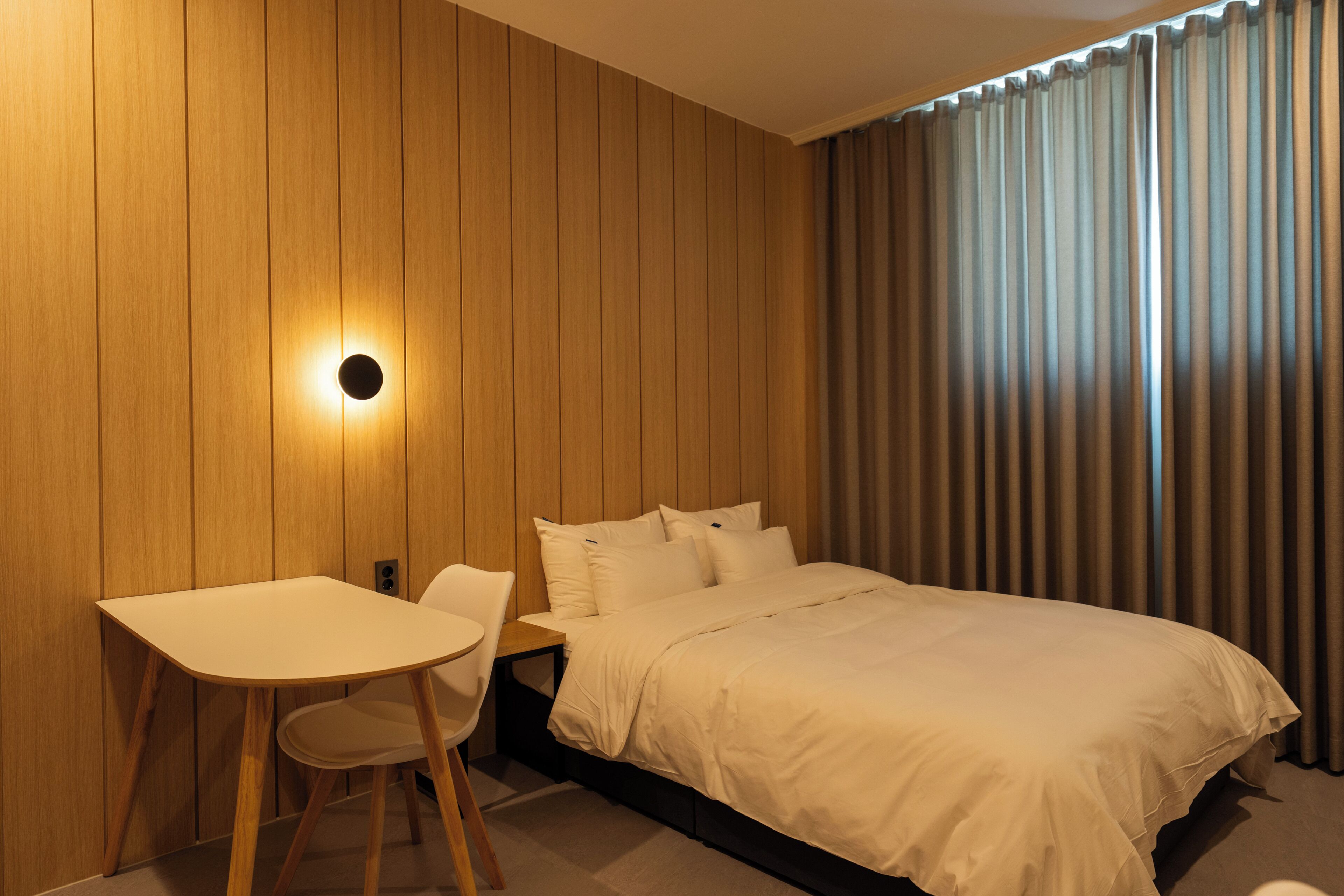 A hotel room with a wooden panel wall, a bed with white bedding, a small table with a chair, and a window with curtains.