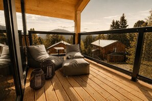 Eine Holzterrasse mit Glastrennwand, gepolsterten Sitzgelegenheiten und Blick auf eine Hütte sowie einen Wald.