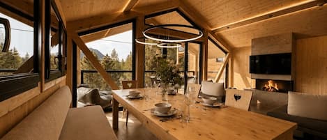 A modern interior with a wooden dining table set for a meal, large windows offering a view of the outdoors, and a fireplace.