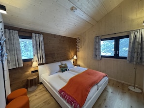 A cozy bedroom with a wooden ceiling, a bed with a colorful pillow, two bedside tables with lamps, and a window with curtains. - Superior Chalet # 56 with Sauna & Hot Tub (Turrach)