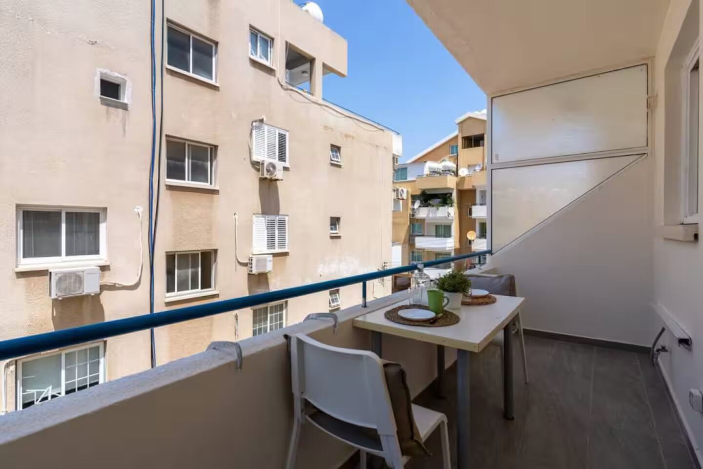 A balcony with a table and chairs, overlooking a residential building with multiple windows and air conditioning units.