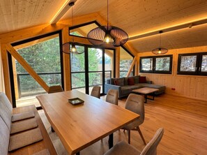 Une pièce spacieuse avec une table et des chaises en bois, un canapé et de grandes fenêtres donnant sur la forêt.