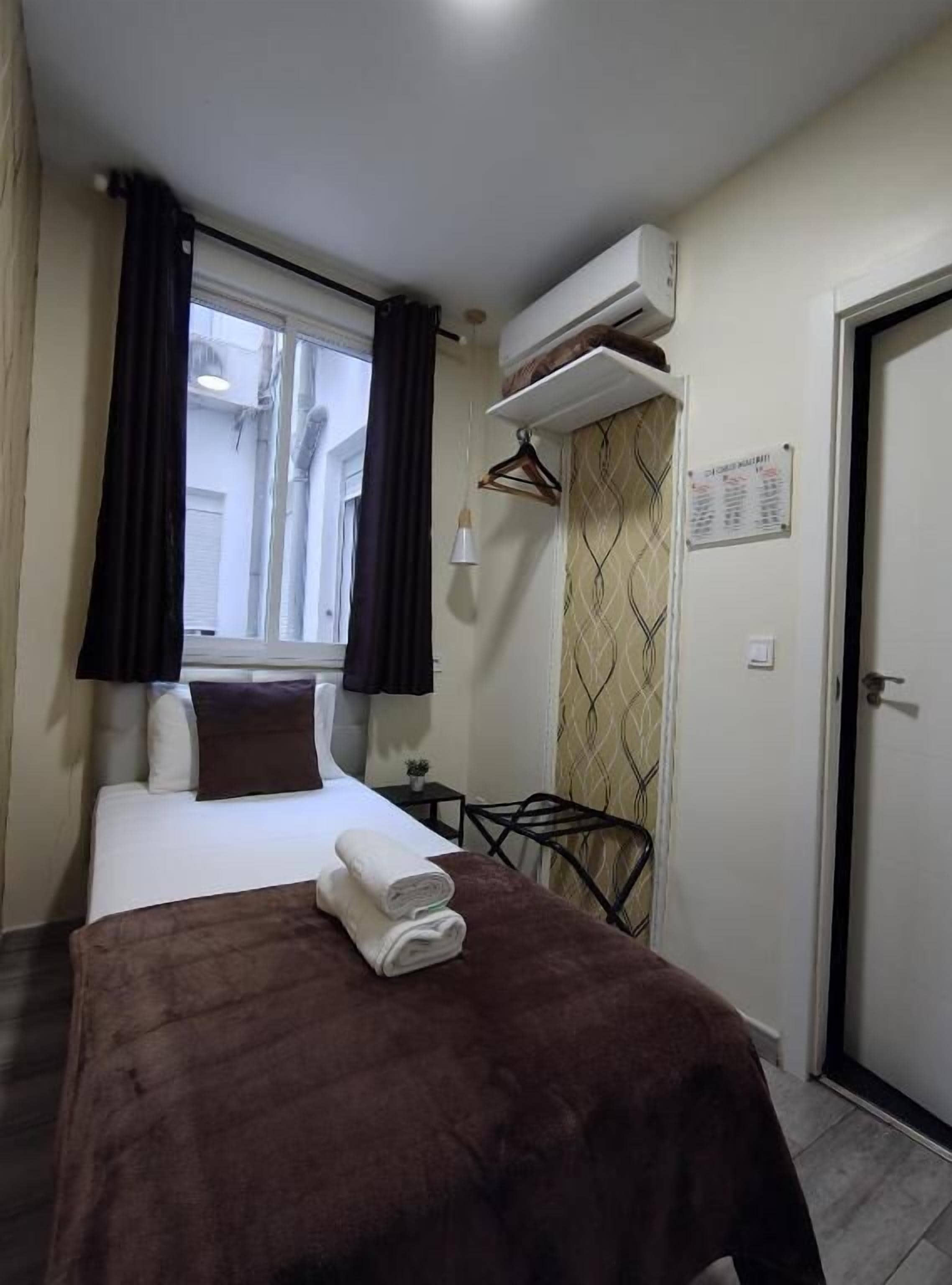 Ein Hotelzimmer mit einem Bett, einem kleinen Tisch, einem Stuhl, einer Klimaanlage, einem Fenster mit Vorhängen und einer Tür.