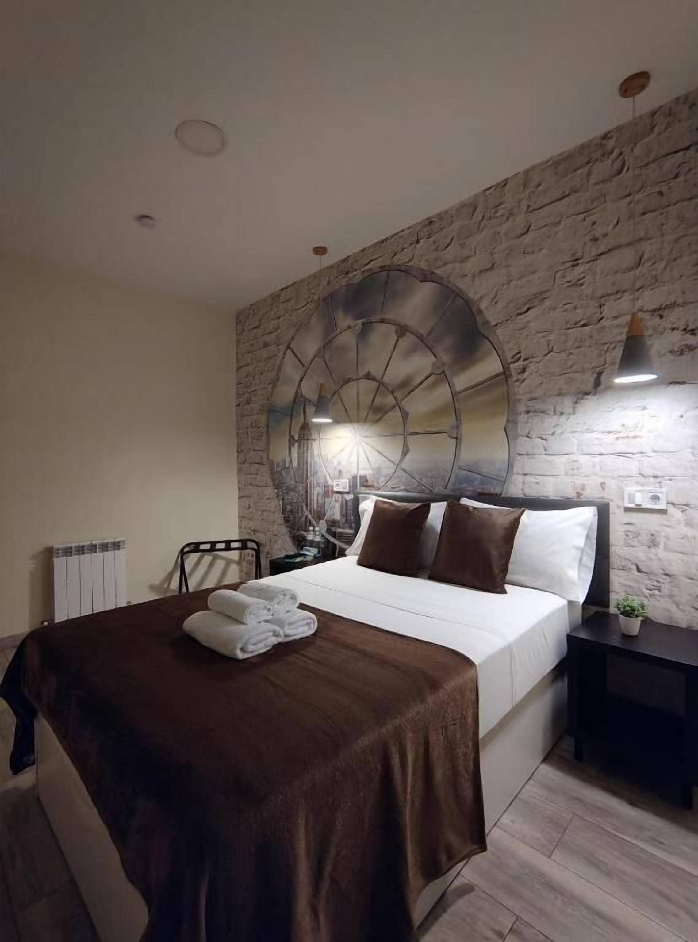 Een slaapkamer met een stenen muur, een grote ronde spiegel, een bed met wit en bruin beddengoed, en een klein nachtkastje met een plant.
