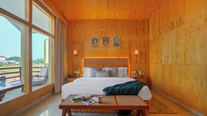 A hotel room with a large bed, a wooden headboard, a window with a view of the outdoors, and a small table with books and a cup. - Manovan Resort by Eastlynn Experiences, Jim Corbett (Kaladhungi)