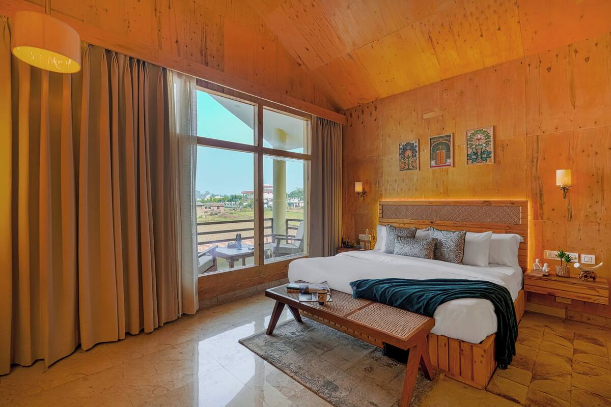 A wooden-finished bedroom with a large bed, a bedside table, a bench, and a view of the outdoors.
