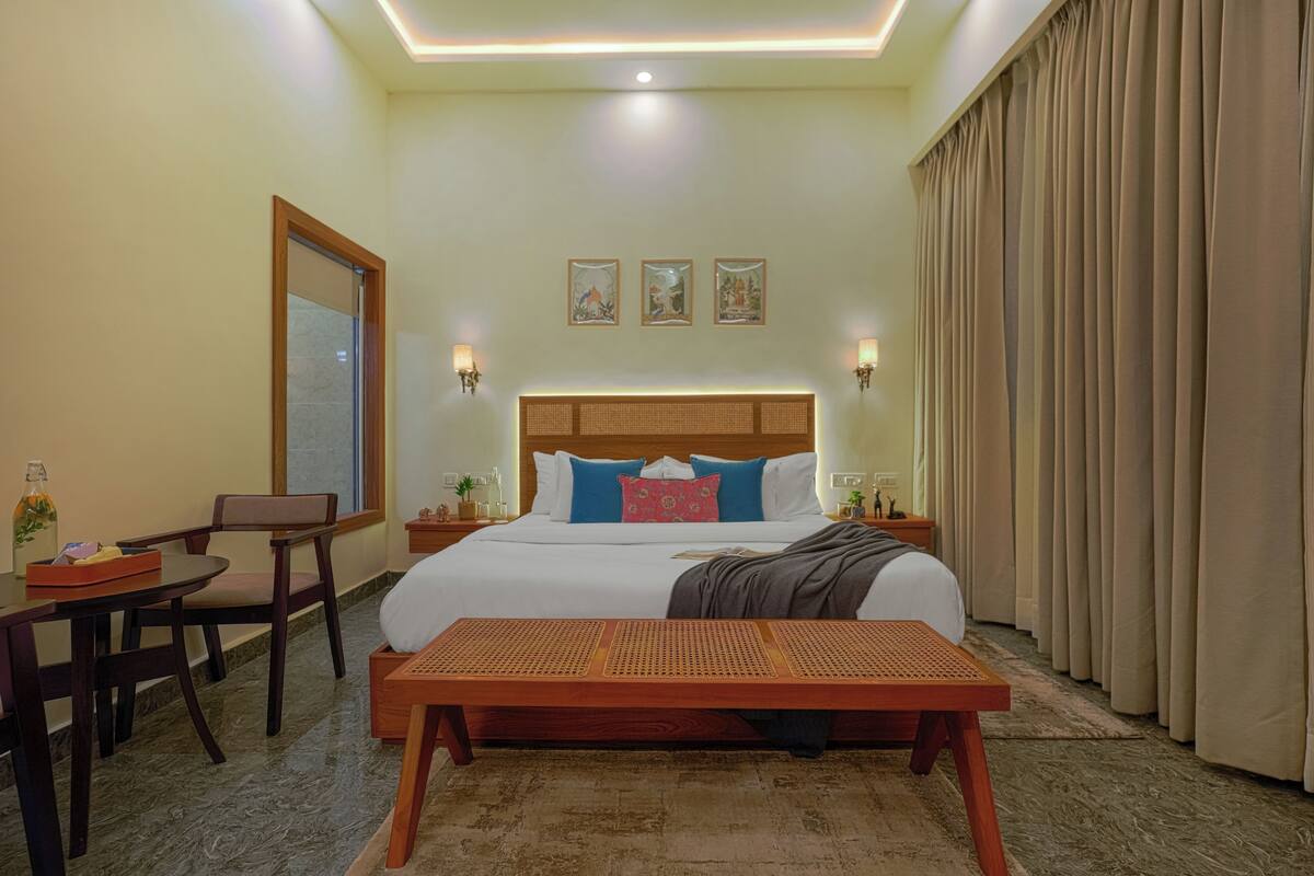 A hotel room with a bed, bedside tables, a desk, chairs, a bench, and curtains.