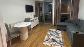 Ein modernes Wohnzimmer mit einem runden Tisch, zwei Stühlen, einem grauen Sofa, einem an der Wand befestigten Fernseher und einer Glastür zum Balkon.