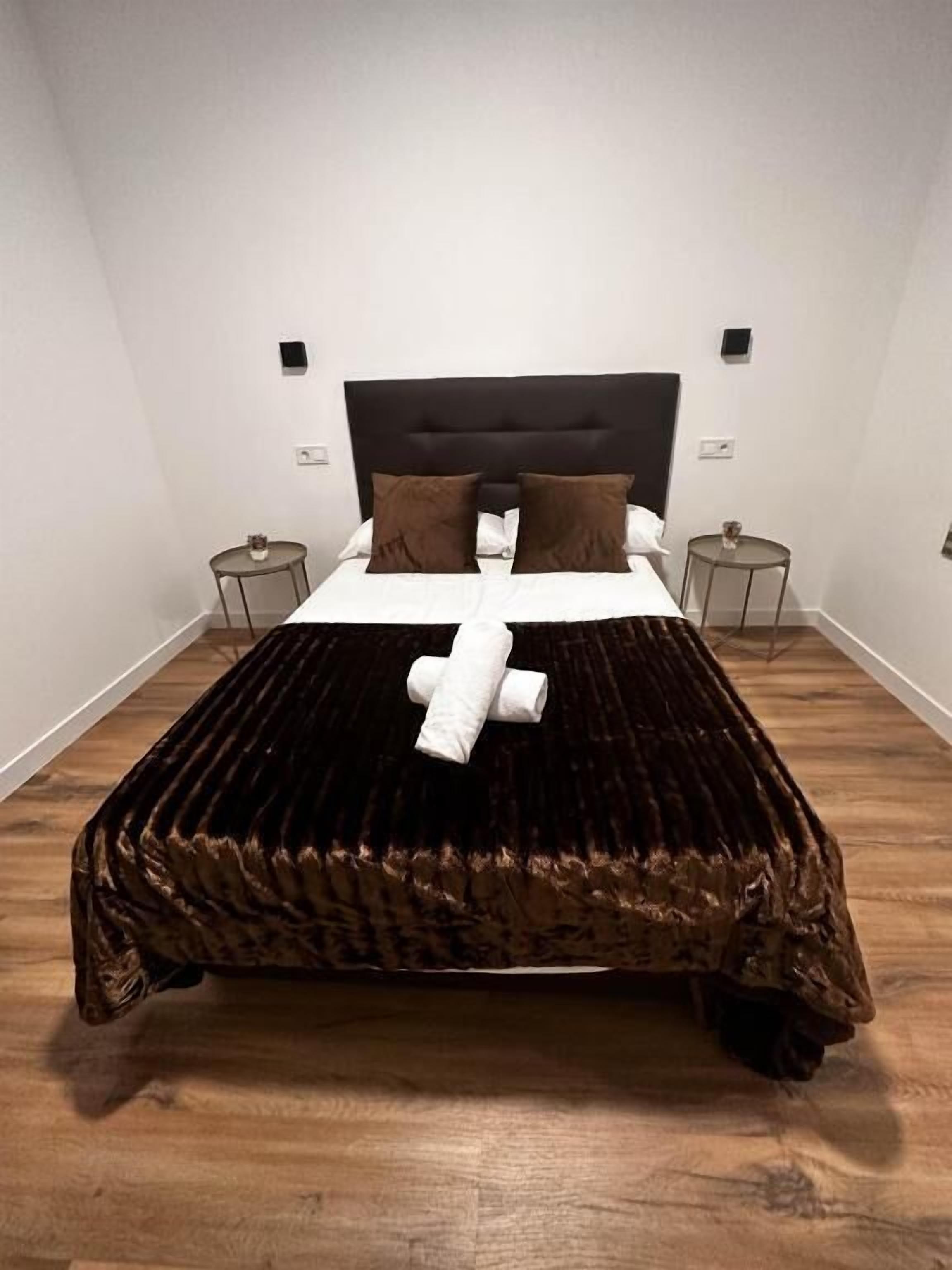 A neatly made bed with a dark brown cover, white linens, and two brown pillows. There are two bedside tables with lamps, and the room has white walls and wooden flooring.
