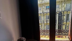 Una habitación con una mesa redonda, un jarrón con flores secas y vistas a un edificio con balcones ornamentales a través de ventanas amplias.