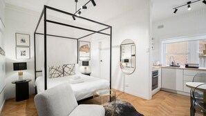 A modern bedroom with a four-poster bed, a sitting area with a chair, a round coffee table, a small side table with a lamp, and a kitchen area with a sink and stove. - Bondi Beachside Escape - Surf & Vibe (Bondi Beach)