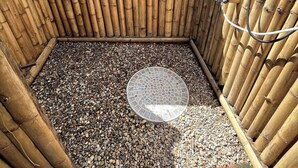 A bamboo-walled outdoor shower area with a circular drain, pebbles on the floor, and a hose on the right. - Boutique Cabin · Peaceful Mountain View (Mueang Ngai)