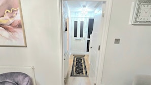 Un salon moderne avec un canapé en cuir, une table à manger dressée avec des assiettes et des couverts, et un couloir menant à une porte surmontée d’un lustre.
