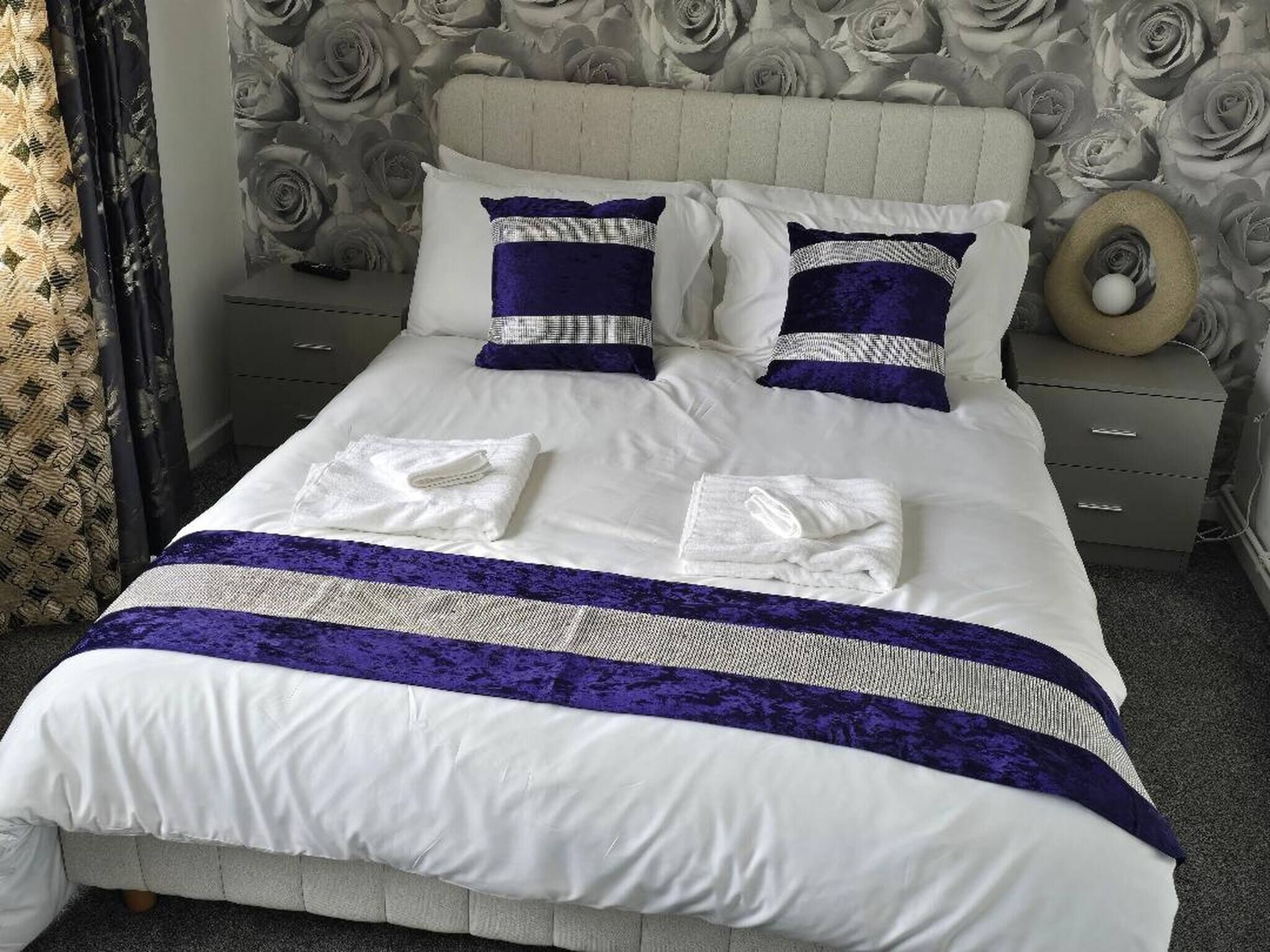 Una cama bien hecha con ropa de cama blanca y morada, dos almohadas y un cabecero decorativo.
