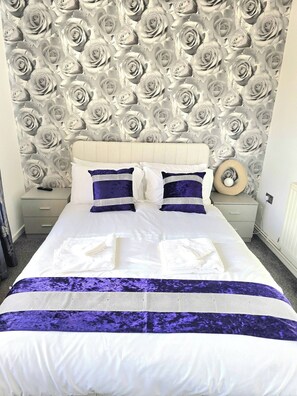 A neatly made bed with white and purple linens, a headboard with a floral pattern, and a bedside table with a lamp. - Gem Place - A home away from home (Hornchurch)