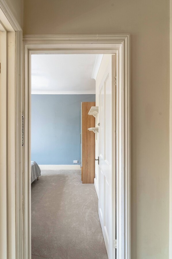 A hallway leading to a bedroom with a bed, a wooden door, and a light switch on the wall. - Elegant 3BR Victorian Retreat, Free Parking (Kingston upon Thames)