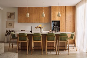 Cocina moderna con armarios de madera, una isla central con sillas verdes, luces colgantes y un florero con flores.