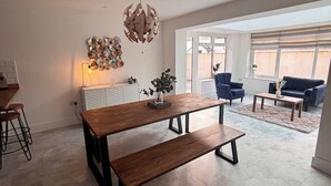 Een ruime woonkamer met een houten eettafel, een bankje en een moderne kroonluchter. Achterin staan twee banken en een bijzettafeltje.
