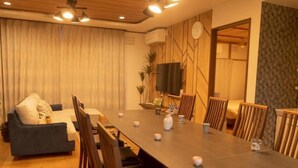 Ein modernes Wohnzimmer mit einer Couch, einem Esstisch und Stühlen. An der Wand ist ein Flachbildfernseher angebracht, außerdem gibt es eine Uhr und eine Pflanze.