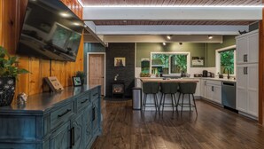 A modern kitchen with a built-in TV, wooden cabinets, a stone fireplace, and a bar area with stools. - Mountain Retreat -Hot Tub -Fire Pit -Stevens Pass -River Access -Pet Friendly (Index)